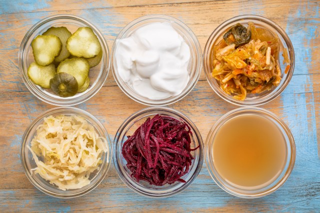 a set of fermented food great for gut health - top view of glass bowls against grunge wood: cucumber pickles, coconut milk yogurt, kimchi, sauerkraut, red beets, apple cider vinegar